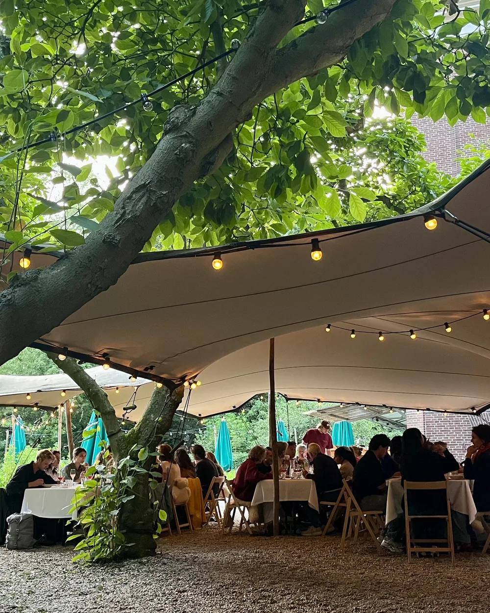 Gasten aan lange tafels onder een tentdoek met lichtjes in de tuin van Huize Frankendael